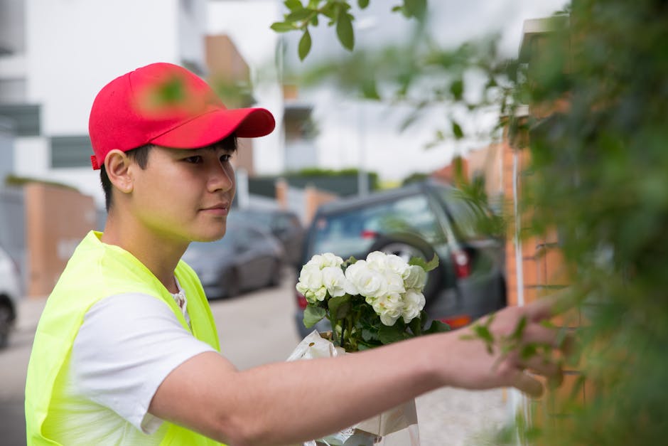 A delivery person wearing a bright pink cap and a white T-shirt is smiling as they extend their hands forward, presenting a bouquet of fresh white roses and greenery. The bouquet is wrapped in clear c