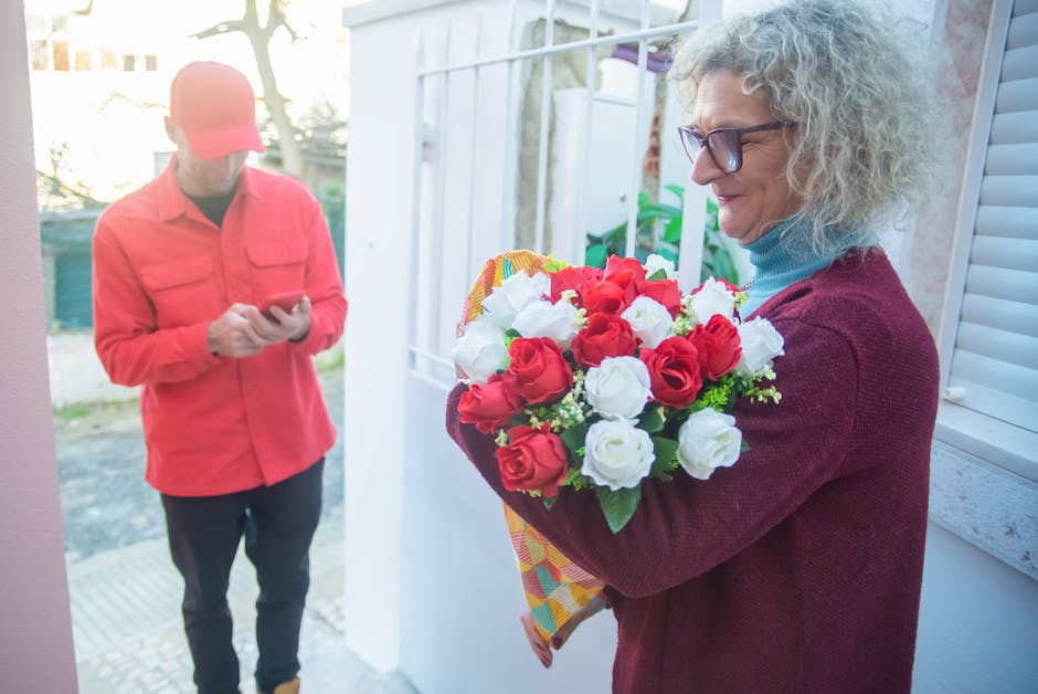 A woman with curly gray hair and glasses is holding a bouquet of fresh red and white roses wrapped in colorful paper, standing near a window with sunlight highlighting her face and the vibrant flowers