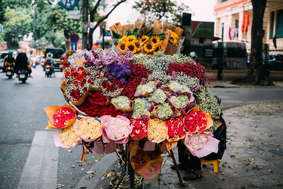 A young man wearing a black helmet navigates a busy street on a motorcycle loaded with large bundles of fresh flowers, primarily pink and green stems, wrapped in brown paper and secured with green tie